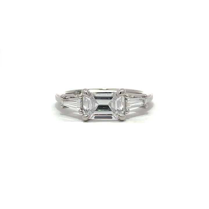  White gold three-stone diamond engagement ring with emerald-cut center stone and baguette side stones on a white background.