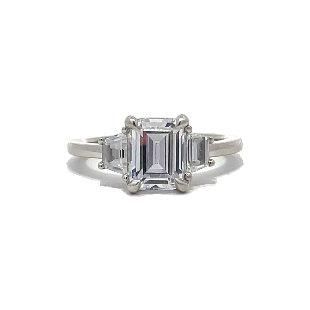  White gold three-stone diamond engagement ring with emerald-cut center stone and baguette side stones on a white background.