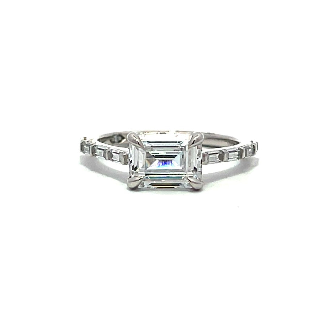 White gold emerald-cut diamond engagement ring with a diamond band on a white background.